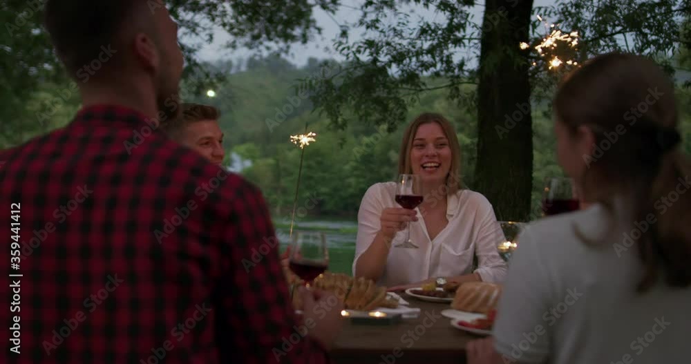 Group of happy friends celebrating holiday vacation using sprinklers and drinking red wine while having picnic french dinner party outdoor near the river Group of happy friends celebrating holiday vacation using sprinklers and drinking red wine while having picnic french dinner party outdoor near the river