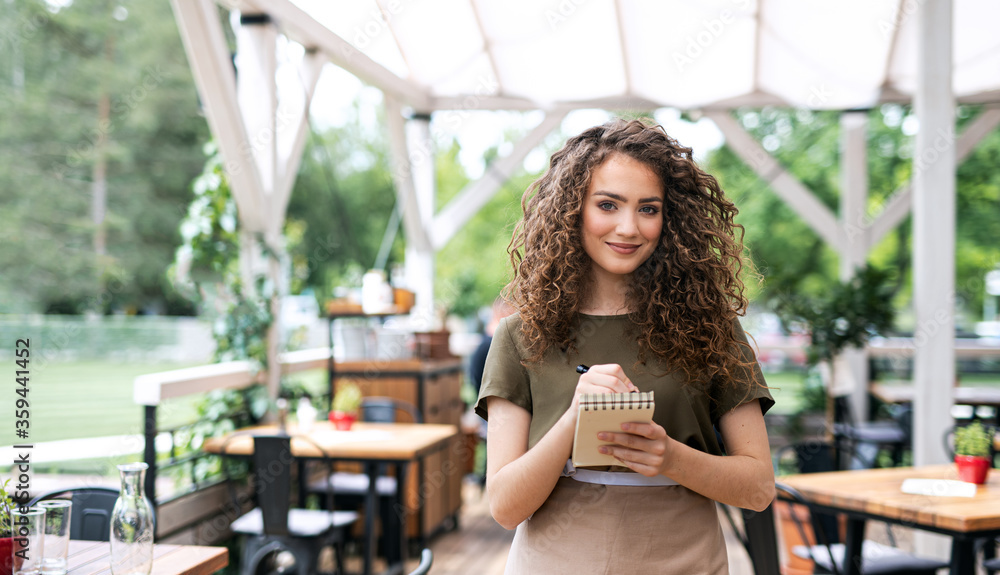 Waitress with order pad standing outdoors on terrace restaurant, looking at camera.
