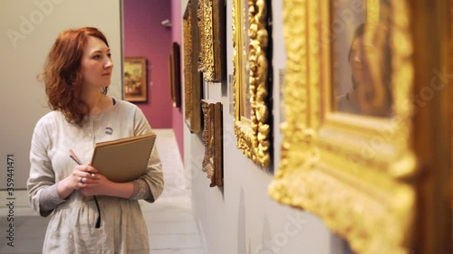 red haired woman in light grey dress with sketchbook walks along wall with hanging pictures in golden vintage frames in light hall of art museum