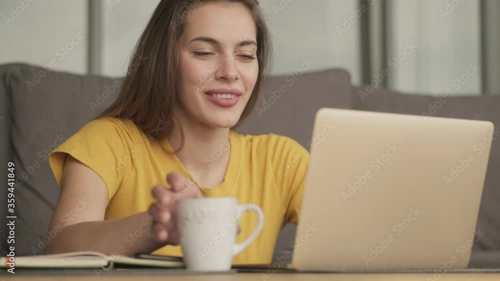A pleased woman is using her laptop while drinking something from a cup while sitting at the table at home A pleased woman is using her laptop while drinking something from a cup while sitting at the table at home