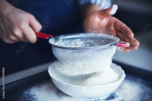 A cook with his hands soiled in flour pours the flour through a red-handled sieve into a white bowl. Cooking process.