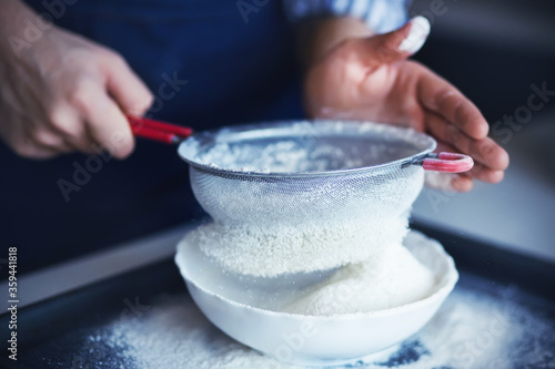 Fotografie A cook with his hands soiled in flour pours the flour through a red-handled sieve into a white bowl