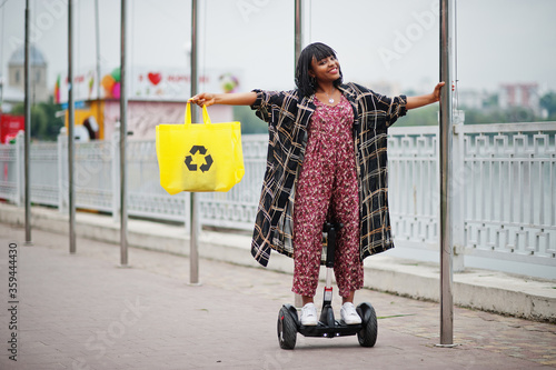 Beautiful african american woman using segway or hoverboard. Black girl with yellow  cloth eco bags recycling symbol. photo