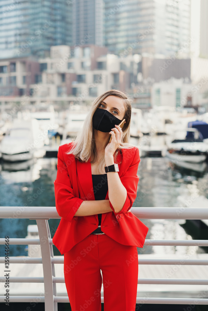 Obraz premium Young businesswoman in red suit and protective face mask standing at city promenade and making a call via a smarphone