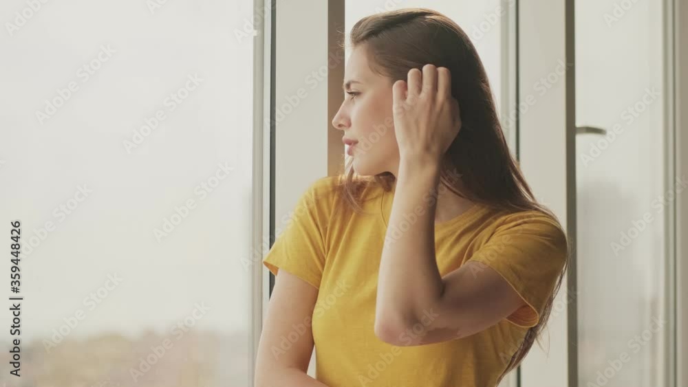 A beautiful young woman is looking out to the window standing in the living room at home A beautiful young woman is looking out to the window standing in the living room at home