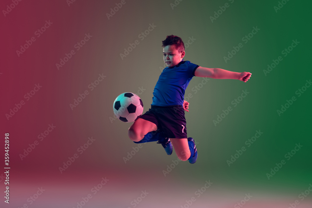 Overcoming. Teen male football or soccer player on gradient background in neon light. Caucasian boy training, practicing on the run, in jump. Concept of sport, competition, winning, motion, action.
