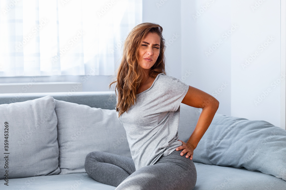 Woman suffering from backache at home. Lower Back Pain . Woman feels back pain massaging aching muscles, sad woman suffers from low-back Woman suffering from backache at home. Lower Back Pain . Woman feels back pain massaging aching muscles, sad woman suffers from low-back