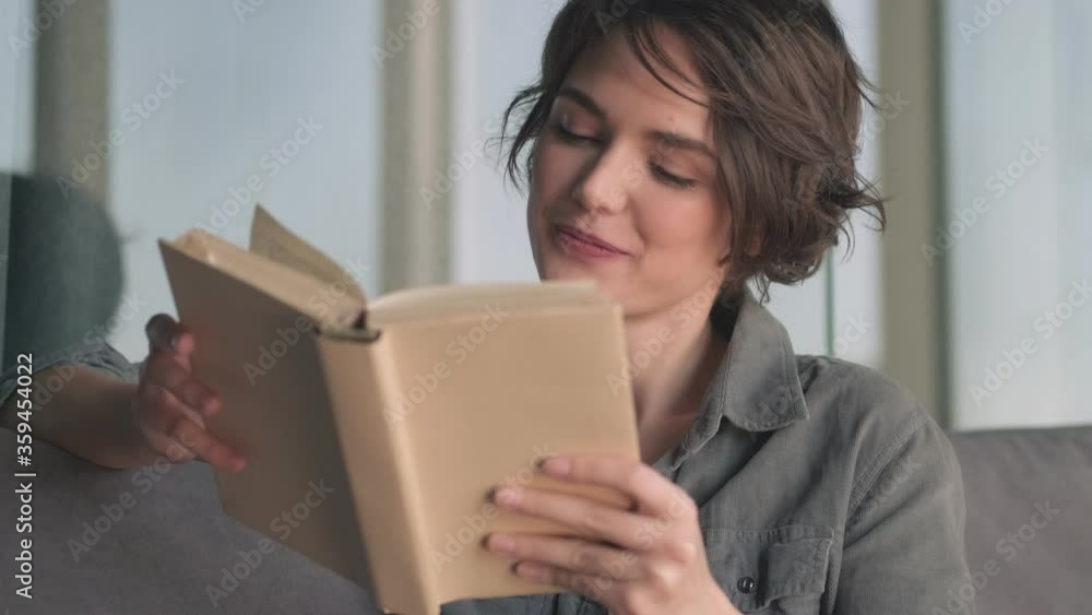 Happy young pretty woman looking away then reading book while sitting on sofa at home Happy young pretty woman looking away then reading book while sitting on sofa at home
