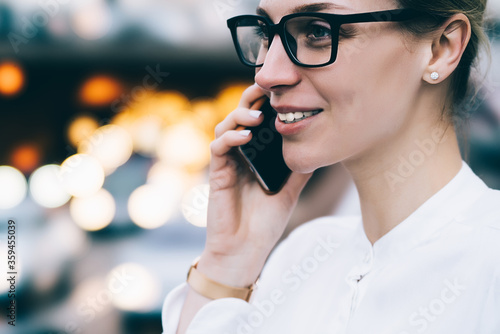 Cropped side view of positive hipster girl laughing while calling on smartphone and talking in roaming on bokeh background.Cheerful young woman in stylish eyeglasses communicating on cellular