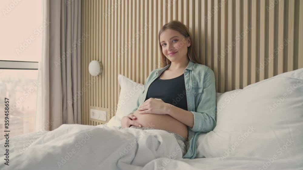 A happy pregnant woman smiling and petting her belly while lying on bed in sunny room A happy pregnant woman smiling and petting her belly while lying on bed in sunny room