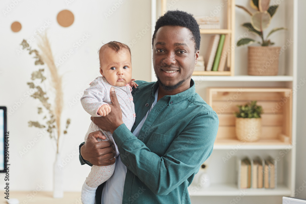 Fototapeta premium Waist up portrait of young African-American man smiling happily at camera while posing with cute baby in home interior, copy space