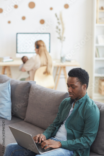 Vertical portrait of modern interracial family working from home, focus on African-American man using laptop in foreground, copy space
