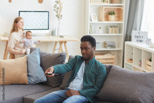 Portrait of young African-American man watching TV at home with woman holding baby in background, copy space
