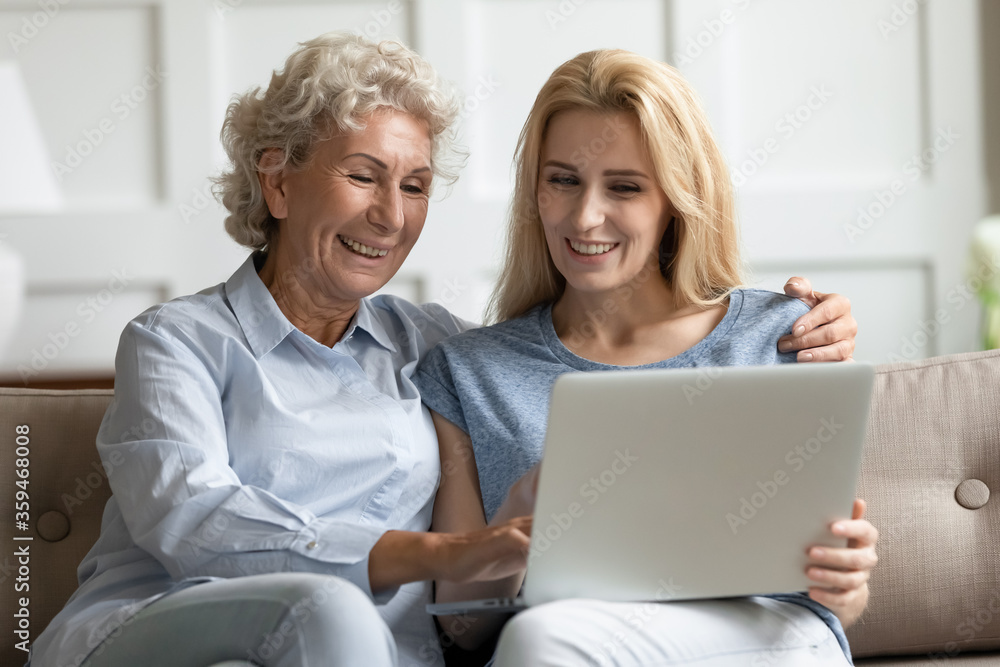 Adult daughter helps to elderly mother with notebook usage, explains teaches older relative how to use device. Family having fun watching movie on computer, buying via internet, store services concept Adult daughter helps to elderly mother with notebook usage, explains teaches older relative how to use device. Family having fun watching movie on computer, buying via internet, store services concept