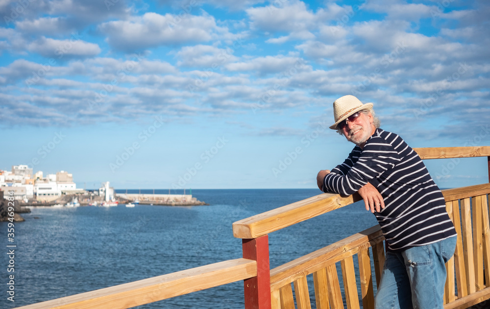 Smiling senior man with straw hat and blue striped shirt in sea excursion, harbour and blue sky on background - active retired elderly people and fun concept