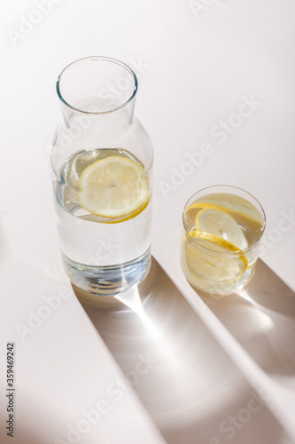 fresh water with lemon slices in carafe and glass on white table with shadows