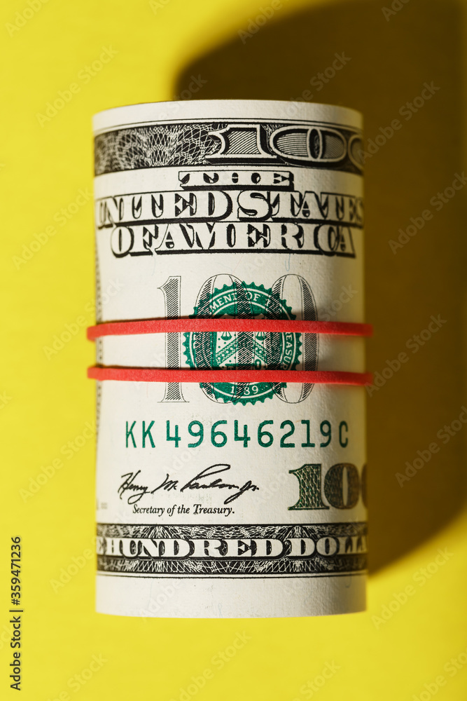 A roll of hundred-dollar American bills is tied with a red elastic band on a yellow background. A roll of hundred-dollar American bills is tied with a red elastic band on a yellow background.
