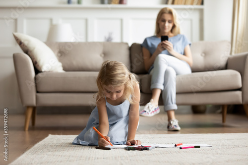 While little daughter drawing with colorful felt-tip pens on album sit on carpet warm floor mom rest on sofa with smartphone gadget device ignoring kid. Weekend leisure, free time, modern home concept