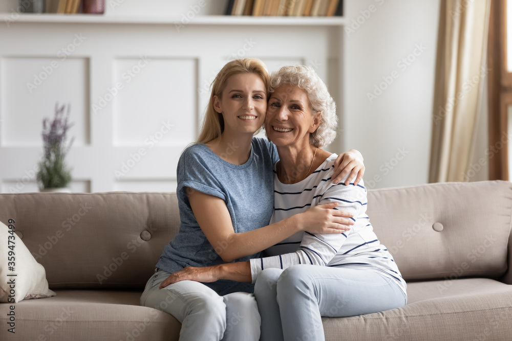 Senior mother hugs young grown up daughter sitting together on couch pose smile look at camera. Happy family portrait, different age and generations loving relatives people, love understating and bond