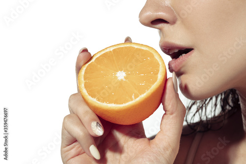 Close up beautiful young woman biting fresh orange slice on white background. Concept of cosmetics, makeup, natural and eco treatment, skin care. Shiny and healthy skin, fashion, healthcare. Copyspace