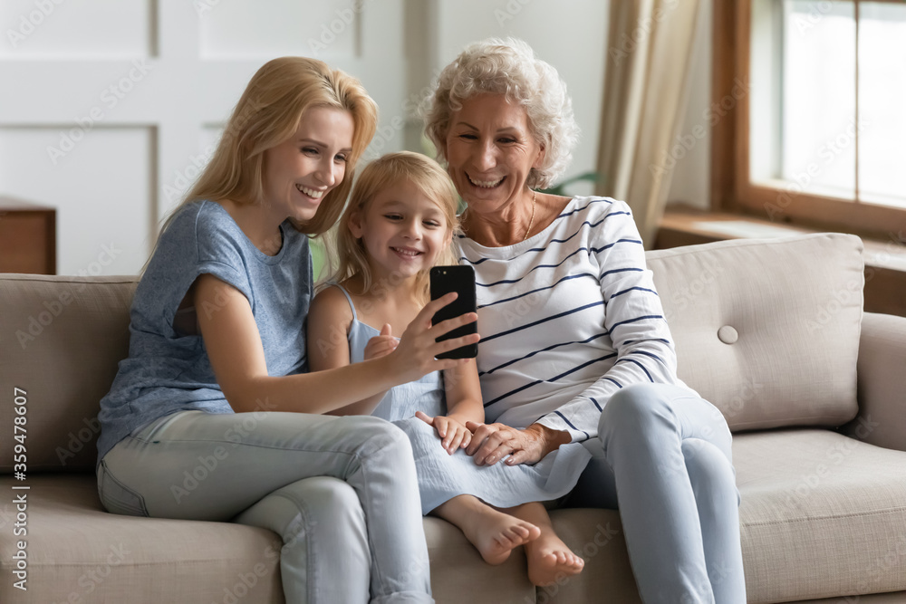 Happy small girl with young mother and aged grandmother at home, multi-generational family sit on sofa in living room use cell, enjoy distant talk via video call, making self-portrait photo for memory