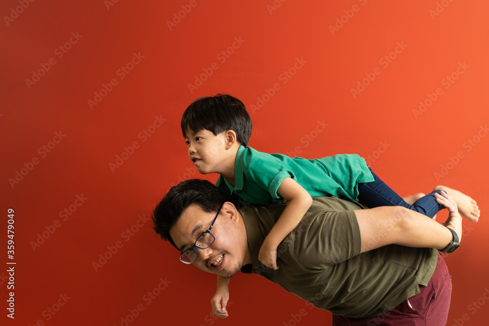 Obraz premium Asian father giving son ride on the back with a red background. Portrait of a happy father giving son piggyback a ride on his shoulders and looking up.