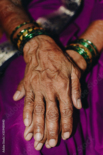 Hands of an elderly indian woman, one on top of the other on her purple saree. Details of the wrinkled brown skin is visible