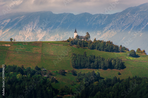 Jamnik church on hilltop with Alps mountains in background. Hill slope covered with grass and trees. Unique perspective of this famous picturesque church in Slovenia