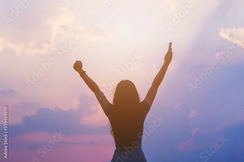 Young empowered lady raising her hands up to the sky, hand holding smart phone against sunset sky photo