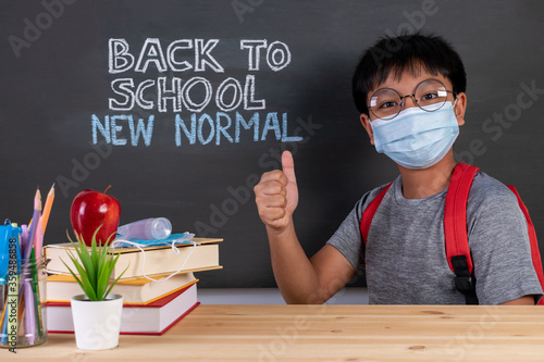 School boy wearing face mask thumbs up over blackboard with text BACK TO SCHOOL NEW NORMAL. Safe back to school during Covid-19 pandemic. New normal education concept.