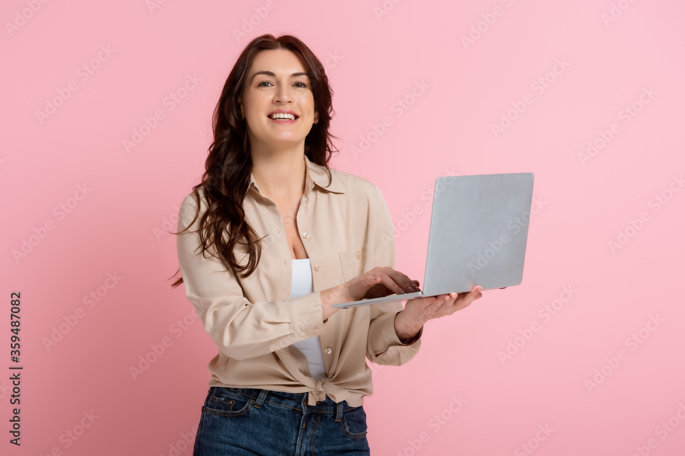 Brunette woman smiling at camera and using laptop on pink background Brunette woman smiling at camera and using laptop on pink background