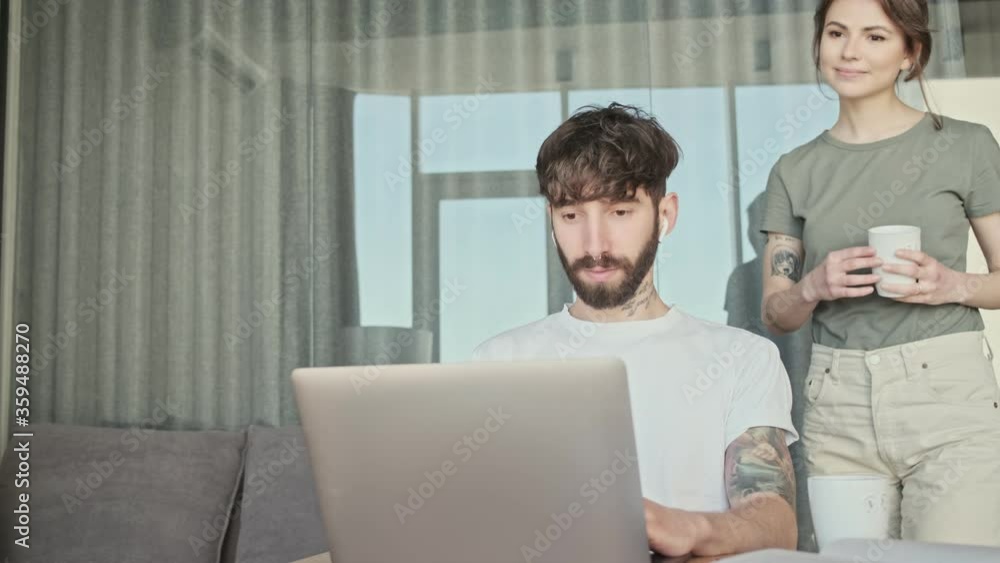 A smiling girlfriend is hugging her boyfriend while he is sitting in the living room and using his laptop at home A smiling girlfriend is hugging her boyfriend while he is sitting in the living room and using his laptop at home