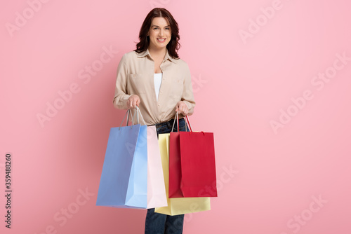 Beautiful woman smiling while holding colorful shopping bags on pink background