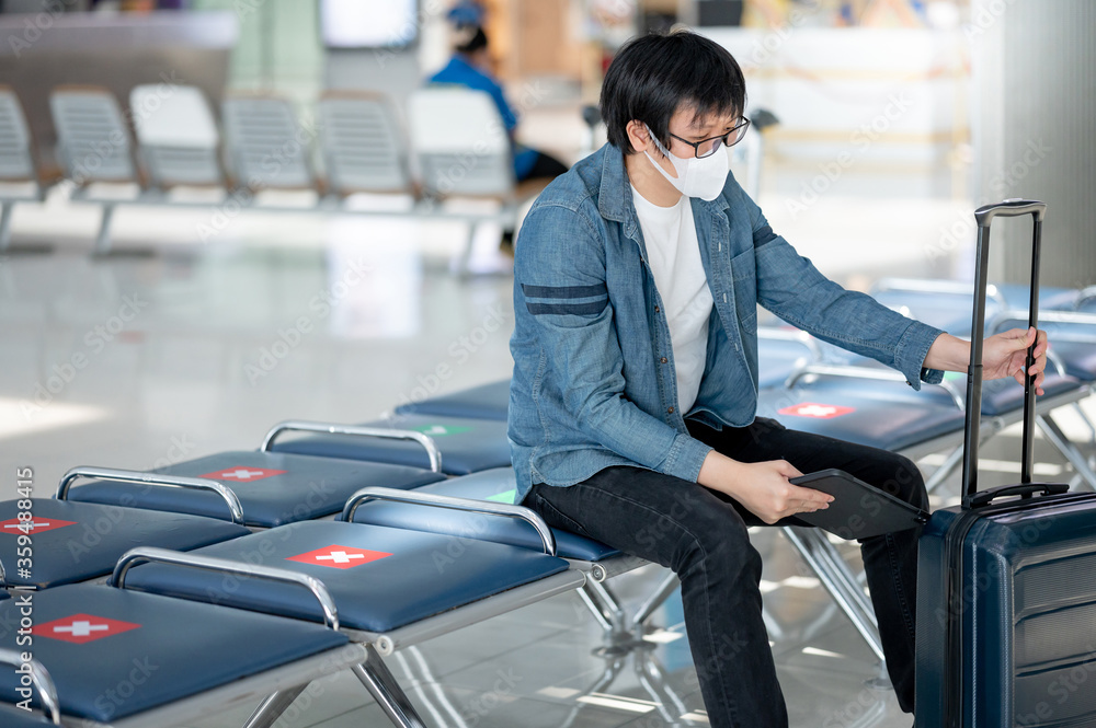 Asian man tourist wearing face mask holding digital tablet and suitcase luggage in airport terminal. Coronavirus (COVID-19) prevention when travel abroad. Health awareness and social distancing
