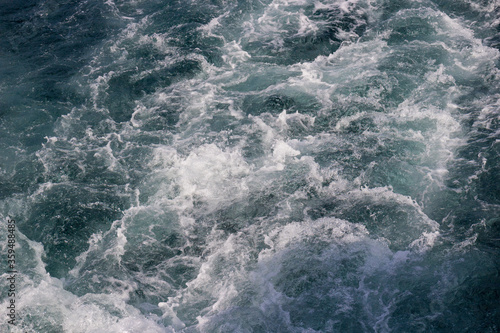 Blurred background of wake water behind a ship on the sea surface. Defocused white foam with shades of blue and green water.