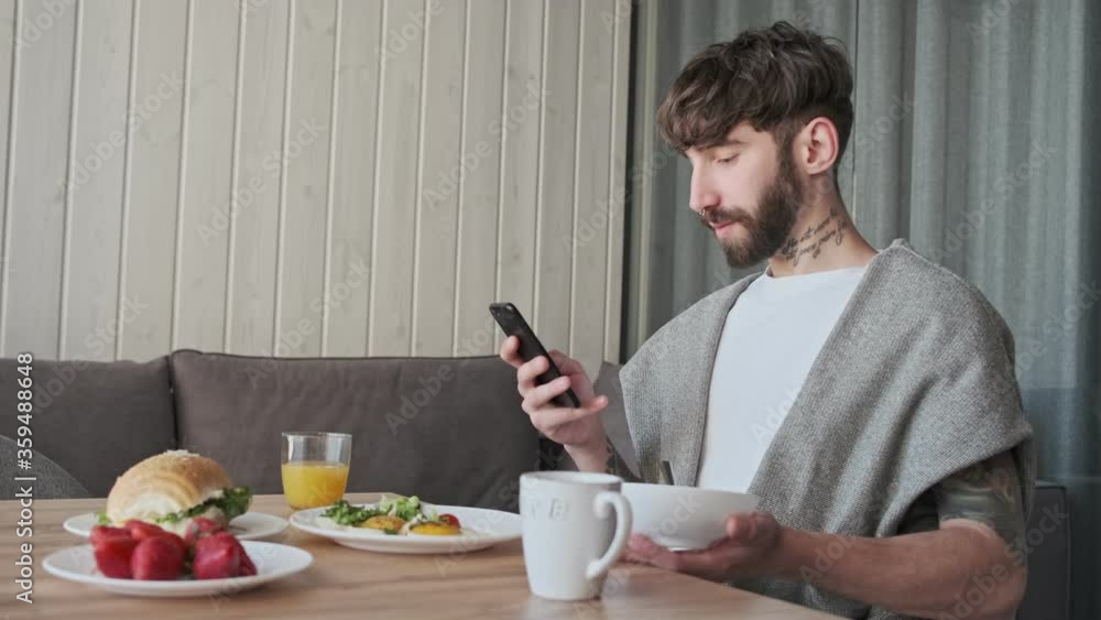 A handsome young hipster man is using his smartphone during breakfast sitting at the table room at home A handsome young hipster man is using his smartphone during breakfast sitting at the table room at home