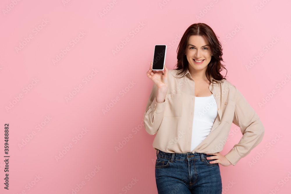 Beautiful woman smiling at camera while showing smartphone with blank screen on pink background Beautiful woman smiling at camera while showing smartphone with blank screen on pink background