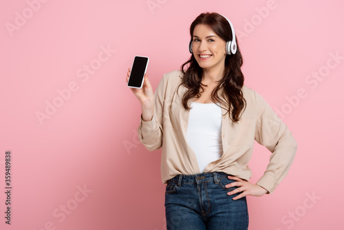 Beautiful woman in headphones holding smartphone and smiling at camera on pink background