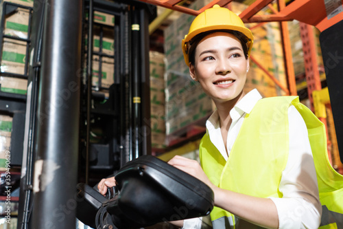 Portrait of Asian warehouse woman worker smile while controlling and driving forklift truck in distribution retail. export import or logistics service business, shipping delivery employee people