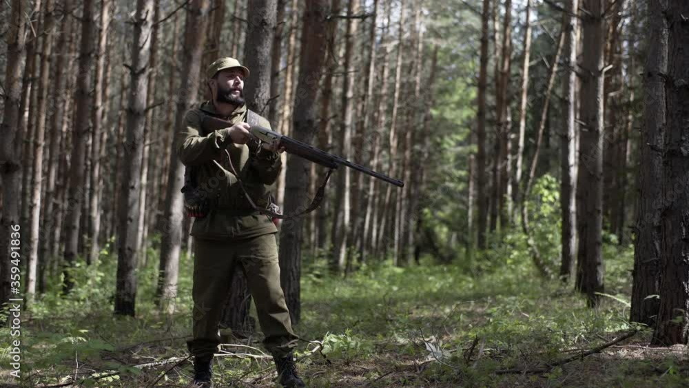 Man takes aim while standing from hunting rifle. Man in comfortable camouflage clothes hunter outdoor in forest hunting alone. Hunter in camouflage aims gun at object in forest during sunset.