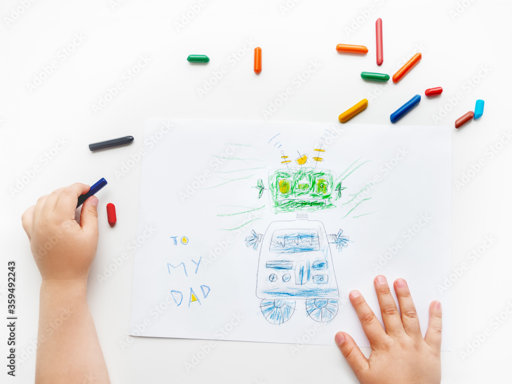 Left-handed toddler draws greeting card with funny robot for Father's day or Daddy's Birthday. Kid uses wax crayons. Top view on child's hands and pencils on white background. Left-handed toddler draws greeting card with funny robot for Father's day or Daddy's Birthday. Kid uses wax crayons. Top view on child's hands and pencils on white background.