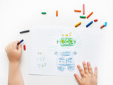 Left-handed toddler draws greeting card with funny robot for Father's day or Daddy's Birthday. Kid uses wax crayons. Top view on child's hands and pencils on white background.