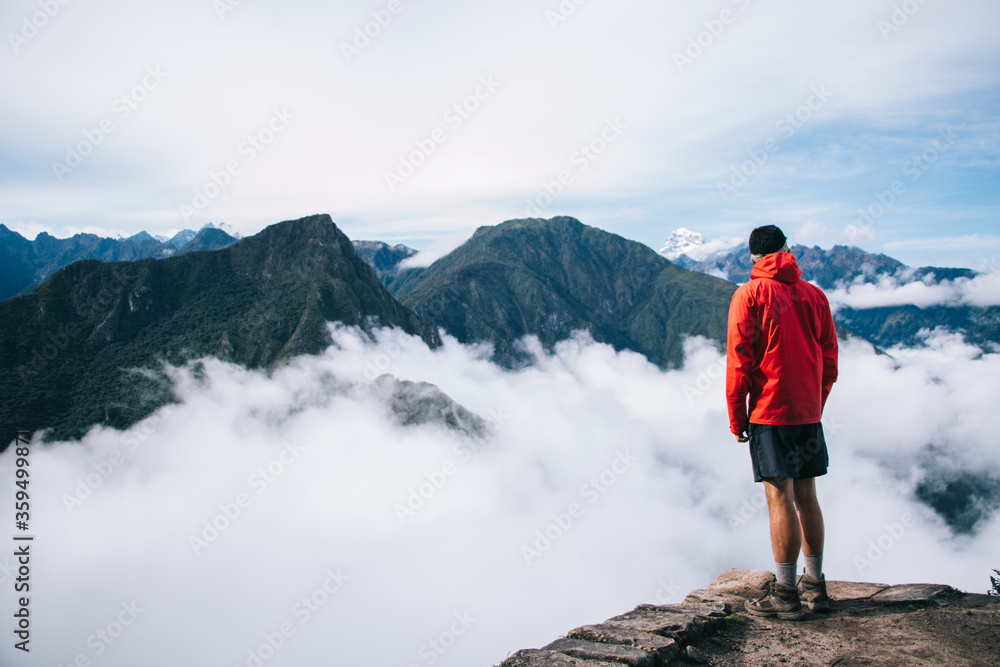 Back view of young man tourist dressed in active wear standing on edge of summit and admiring breathtaking view of green mountains covered white mist.traveler enjoying natural beauty of environment Back view of young man tourist dressed in active wear standing on edge of summit and admiring breathtaking view of green mountains covered white mist.traveler enjoying natural beauty of environment