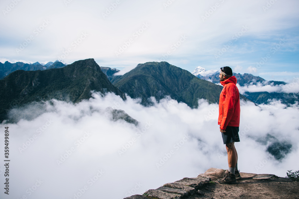 Experienced male tourist dressed in active wear enjoying calm and inspiration of high green mountains covered white fog standing on edge of summit.Young man traveler admiring breathtaking view