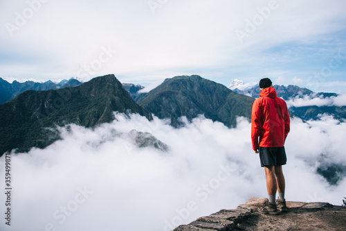 Back view of young man tourist dressed in active wear standing on edge of summit and admiring breathtaking view of green mountains covered white mist.traveler enjoying natural beauty of environment