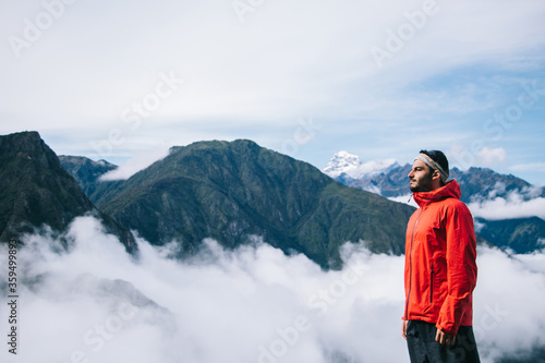 Side view of young bearded man dressed in active wear for trip standing on background with high mountains covered mist during trekking wanderlust.Male traveler admiring beauty of natural environment