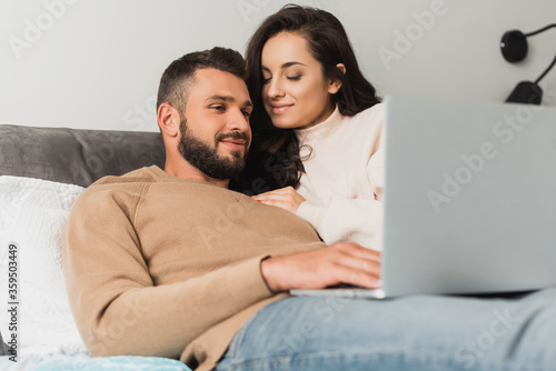 selective focus of happy freelancer using laptop near beautiful girlfriend in bedroom