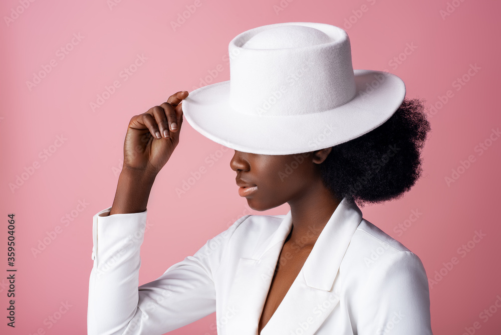 Elegant beautiful African American woman wearing classic white blazer, hiding eyes by stylish hat, posing in studio, on pink background. Close up profile portrait. Elegant beautiful African American woman wearing classic white blazer, hiding eyes by stylish hat, posing in studio, on pink background. Close up profile portrait.
