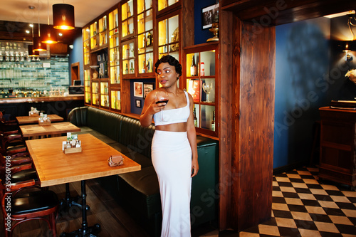 African american woman, retro hairstyle in white dress at restaurant with glass of wine.