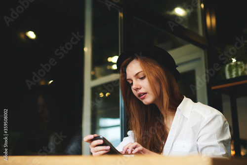 portrait of well dressed female beauty blogger posting publication and multimedia files in social networks waiting for feedback from followers spending leisure in cafe using free internet connection